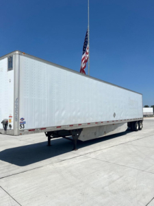 Utility van trailer shown in side view with a white corrugated aluminum exterior, dual rear axles, landing gear, and a flag in the background.