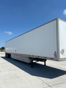 Utility van trailer side view showing a long white semi-trailer with reflective tape and tandem axle underside.