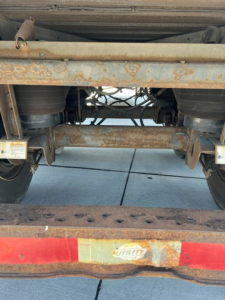 Trailer rear undercarriage showing axles and leaf springs with rust. Utility brand plate on rear bumper visible.