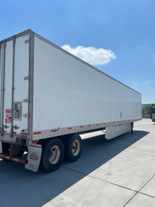 Utility van trailer showing side and rear with tandem axles and plain white corrugated metal exterior.