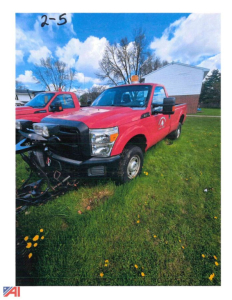 Ford F-350 Super Duty pickup with red V-plow attachment, front view in grassy lot, side badge visible, blue sky with clouds.