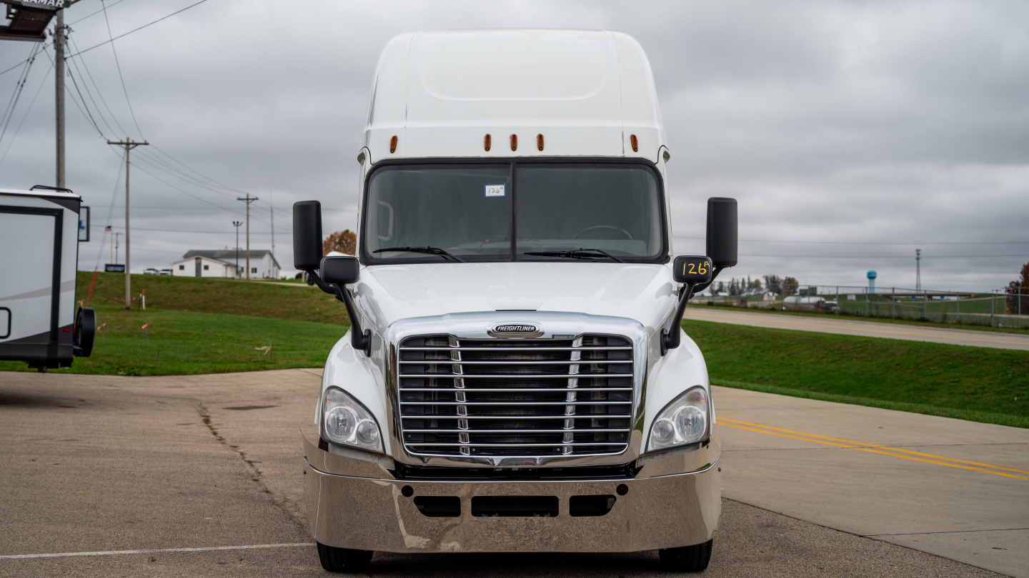 Truck front view showing chrome grille and Freightliner emblem on a white sleeper cab tractor.