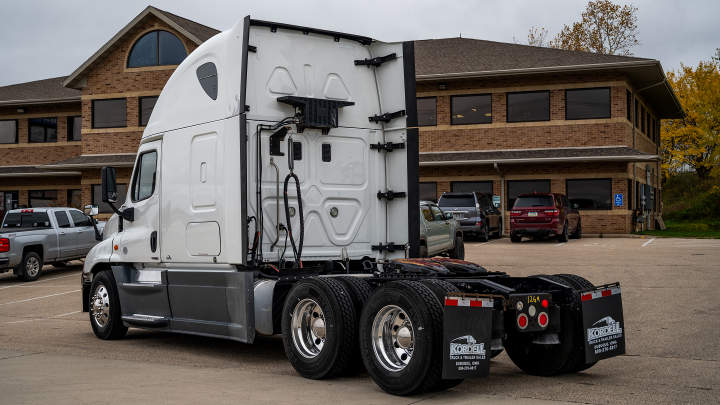 Truck cab and rear view showing Freightliner Cascadia 125 semi-truck tractor, side profile, tandem rear axles, in a parking lot with building in background.