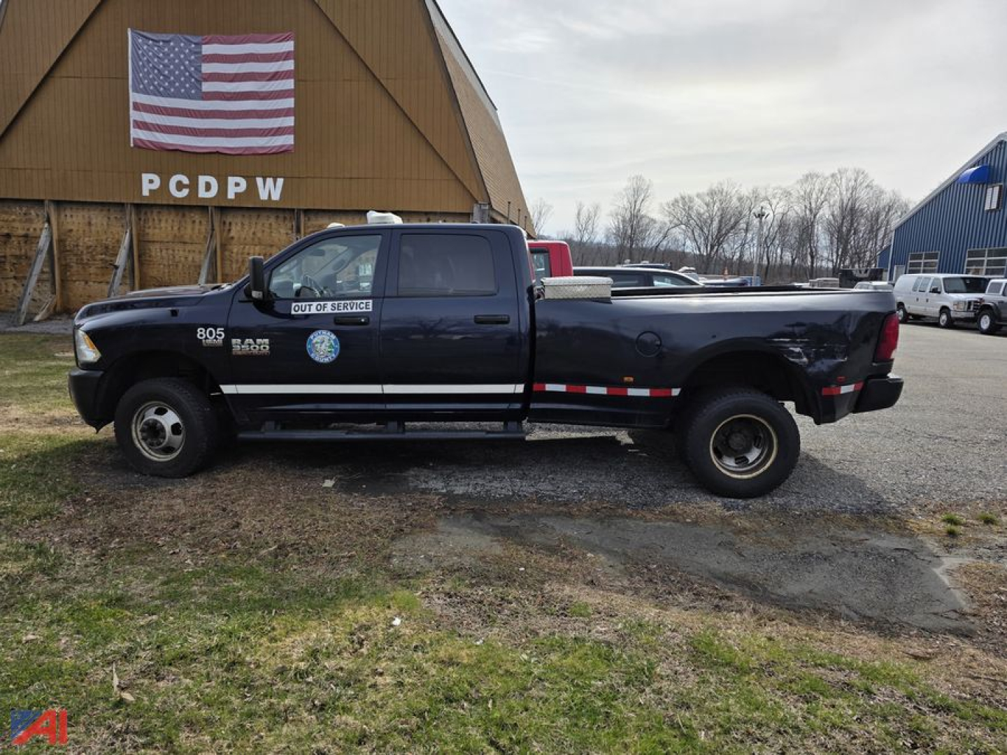 2017 Ram 3500 pickup truck in dark blue with extended cab, service decals on door, large white letters 'OUT OF SERVICE' on the front passenger door, and a red/white/blue flag visible. The truck is parked on a grassy area near a building wit