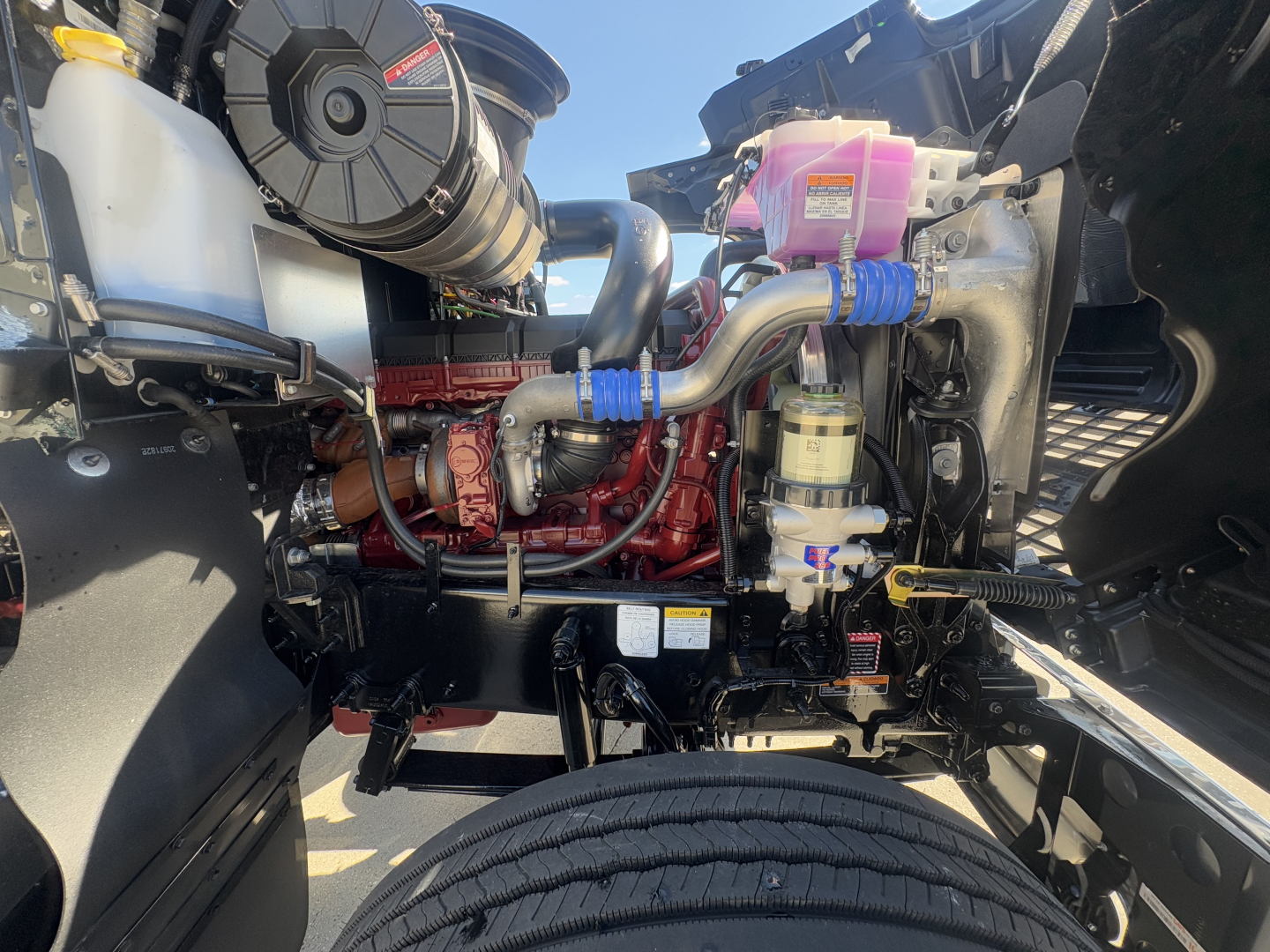 Mack PI64T engine bay showing coolant reservoir, pink coolant overflow bottle, blue hose clamps, red engine block, air intake, and various hoses in foreground.