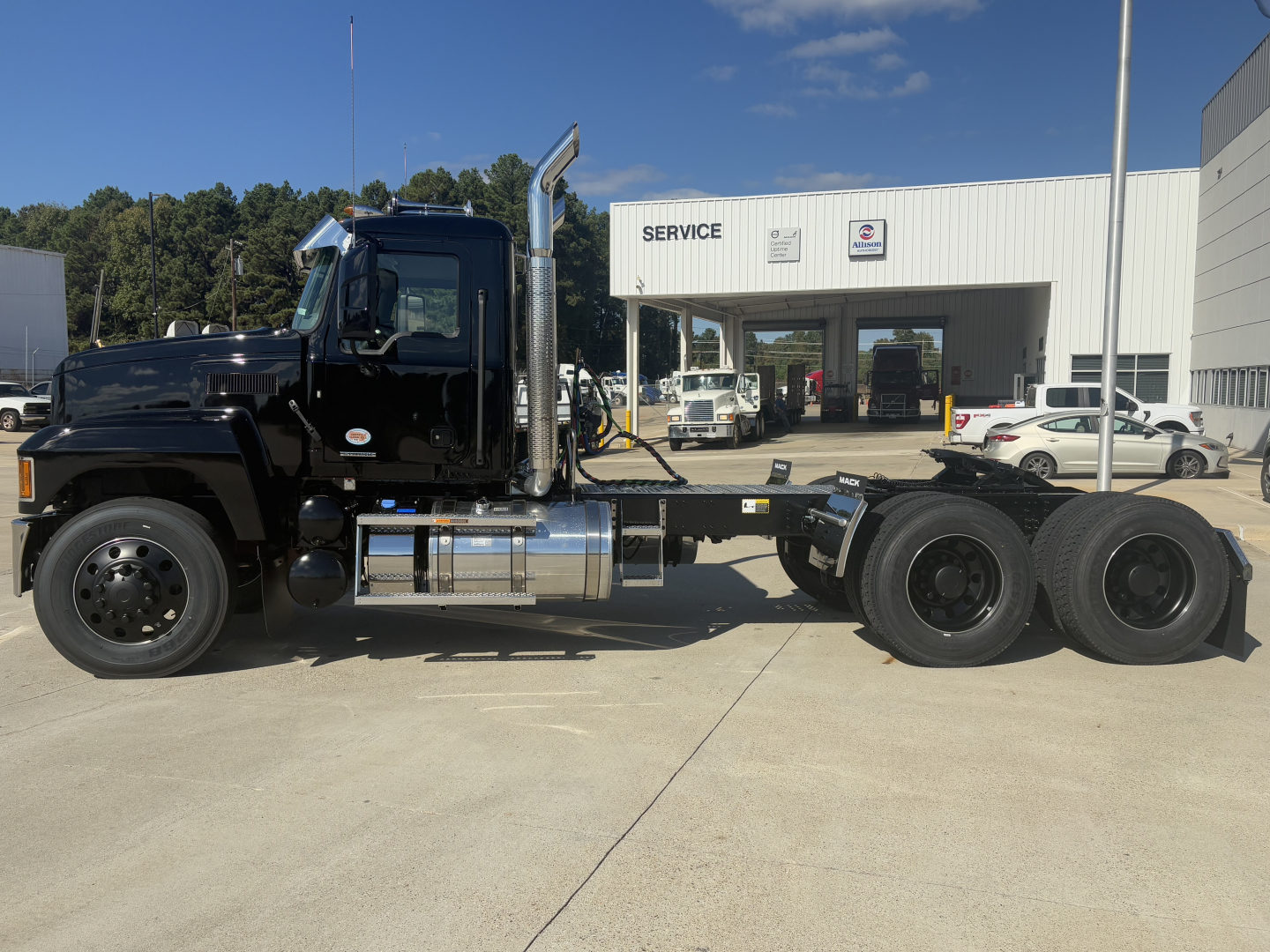Truck chassis and cab side view showing Mack PI64T with black cab, chrome exhaust stack, fuel tanks, and three rear axles. Visible text includes SERVICE on building in background.