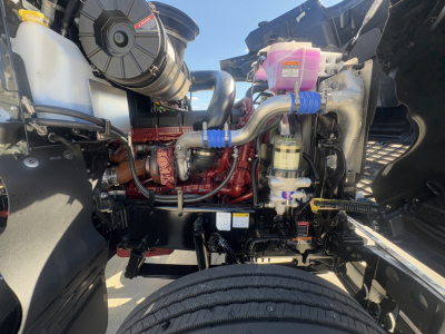 Mack PI64T engine bay showing coolant reservoir, pink coolant overflow bottle, blue hose clamps, red engine block, air intake, and various hoses in foreground.