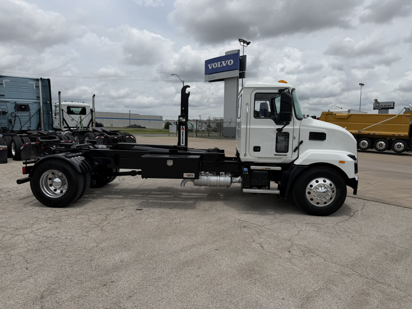 Side view of a white 2024 Mack MD6 hooklift truck chassis with empty frame, black rails, and chrome wheels. Visible text includes a Volvo sign in background.