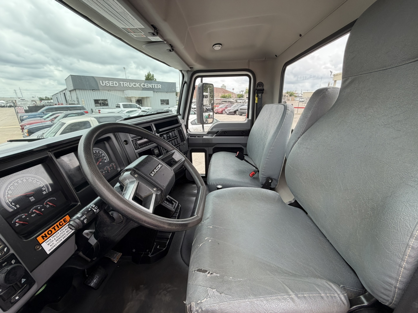 Cab interior of a 2024 Mack MD6 hooklift truck showing steering wheel, instrument cluster, driver and passenger seats with wear on seat edge.