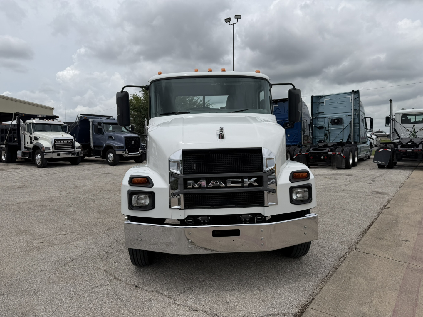 Mack MD6 front view showing large grille with MACK emblem, white cab, chrome bumper, and clearance lights; truck in a lot with other trucks.