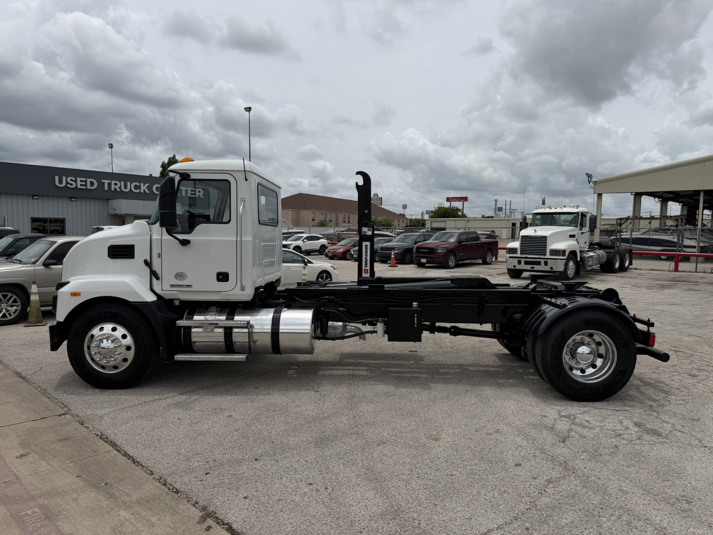 White Mack MD6 hooklift truck chassis side view showing fuel tank, exhaust, and hydraulic components.