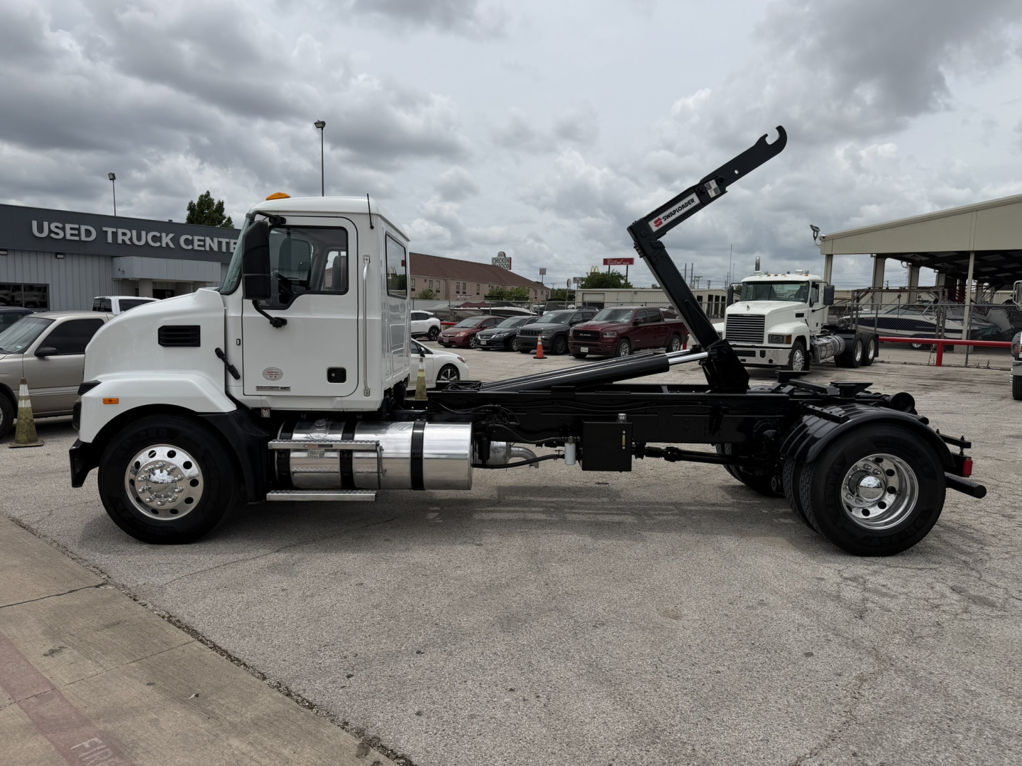 Truck chassis with hooklift system visible; cab shows chrome fuel tanks and exhaust stack; rear area includes hydraulic hooklift arm and frame; background shows dealership building.