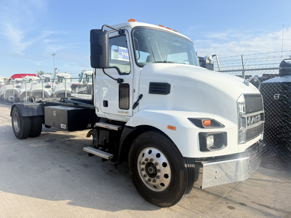 White Mack MD6 hooklift truck from side/front-left angle showing cab, front bumper, wheel, and chassis; no visible numbers readable except potential stickers on door.