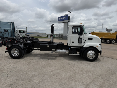 Side view of a white 2024 Mack MD6 hooklift truck chassis with empty frame, black rails, and chrome wheels. Visible text includes a Volvo sign in background.