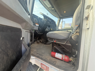 Truck cab interior showing steering wheel, dashboard, driver seat, and a red fire extinguisher on the floor; visible text includes stickers and the VIN plate area.