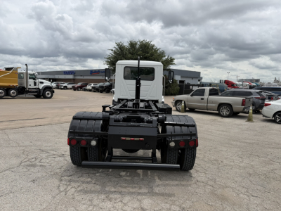 Rear view of a white cab Mack MD6 tractor with black fifth wheel and dual rear axles in a lot; distant vehicles and dealership buildings visible.