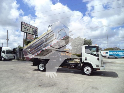 Isuzu NPR HD dump truck in a dealership lot with its dump bed raised. Background shows signs and other trucks.