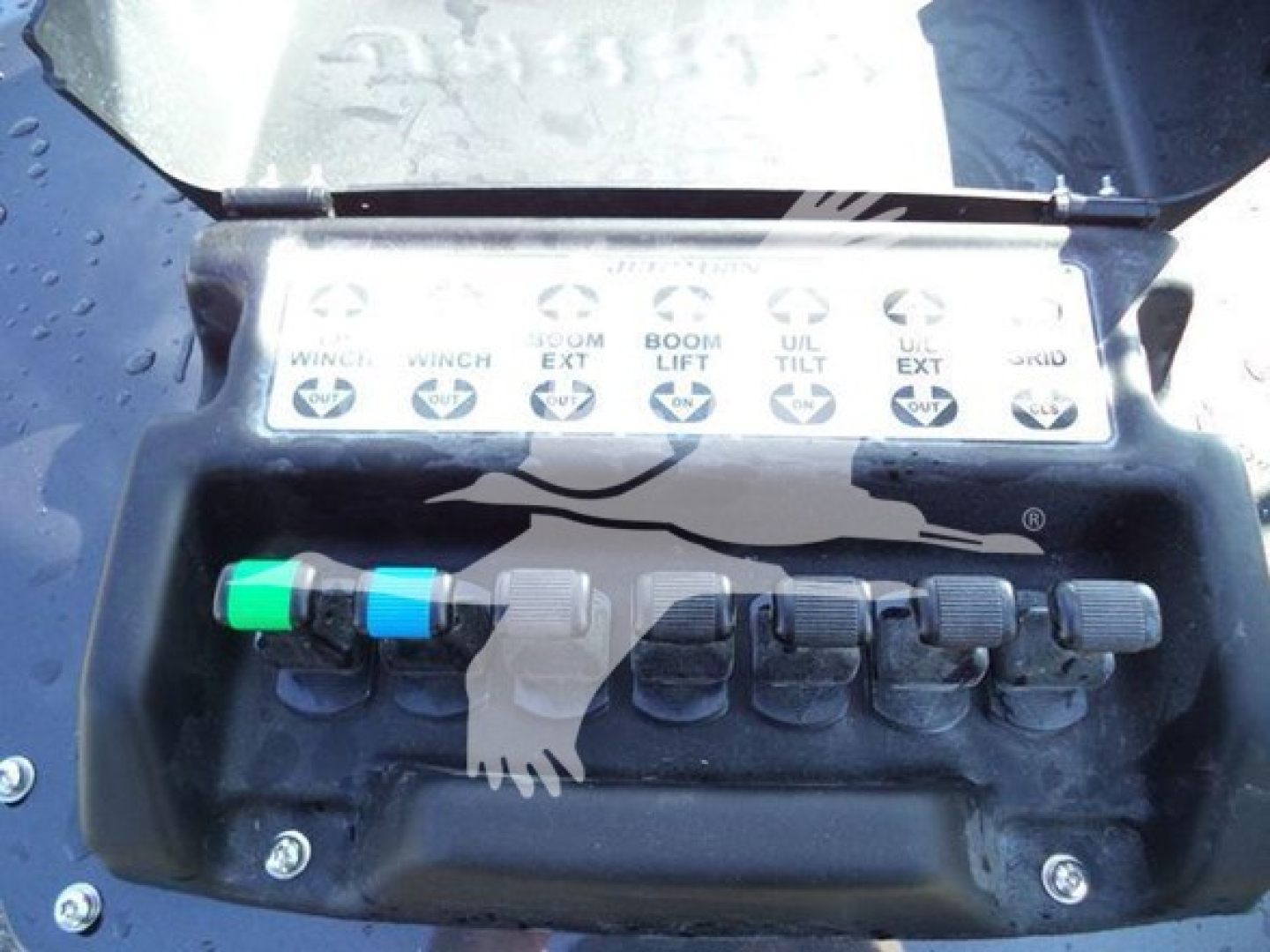 Control panel with labeled levers for winch operations mounted on a truck winch; labels read from left: 'WINCH', 'WINCH', 'SLOW EXT', 'BOOM LIFT', 'TILT', 'UL', 'UP', etc. There are colored buttons/levers (green, blue) and multiple black kn