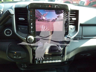 Dashboard view of a Ram truck interior with a large touchscreen display showing a rear camera image. Visible controls include circular dials on both sides, climate controls, and a row of toggle switches below. The overlay shows a ghostly ha