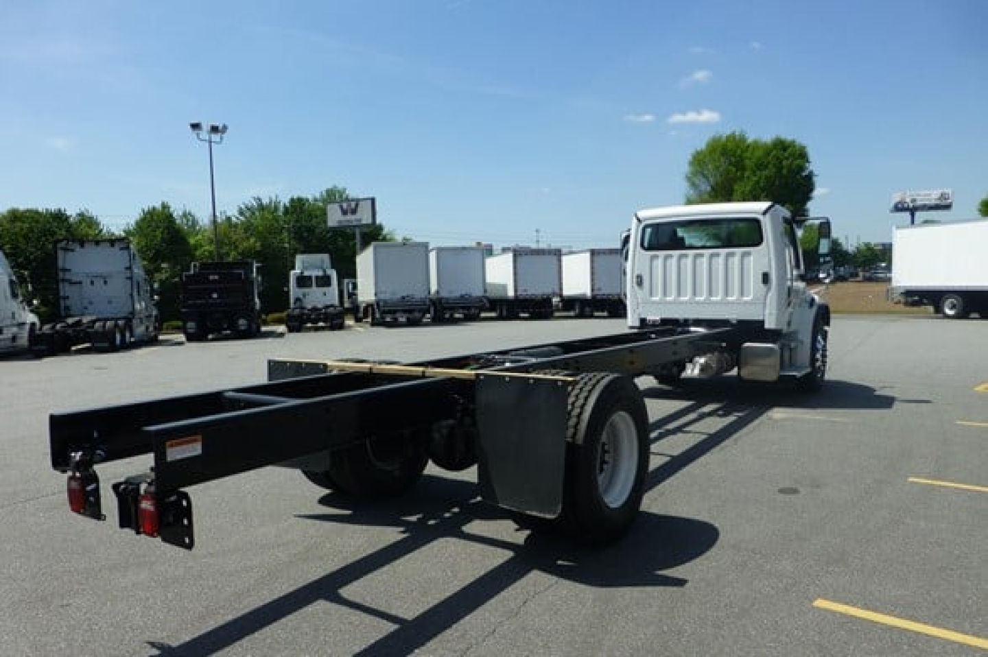 Freightliner Business Class M2 106 Plus cab and chassis frame seen from rear-left side, with white cab and long black chassis frame; trailer connections and rear lights visible