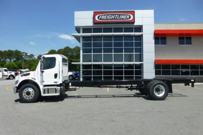 Freightliner Business Class M2 106 Plus cab and chassis in front of dealership showing white cab and long bare chassis frame.