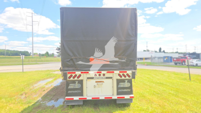 Rear view of a Benson flatbed trailer with a dark tarps cover, showing suspension and bumper, license/branding area with Conestoga logos partially visible.