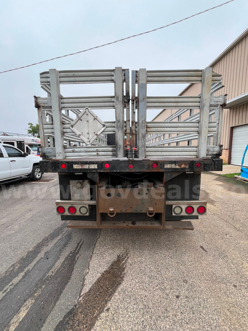 Back of flatbed dump truck with metal gating and dual taillights. Visible text includes a diamond placard holder on the gate and taillight cluster.