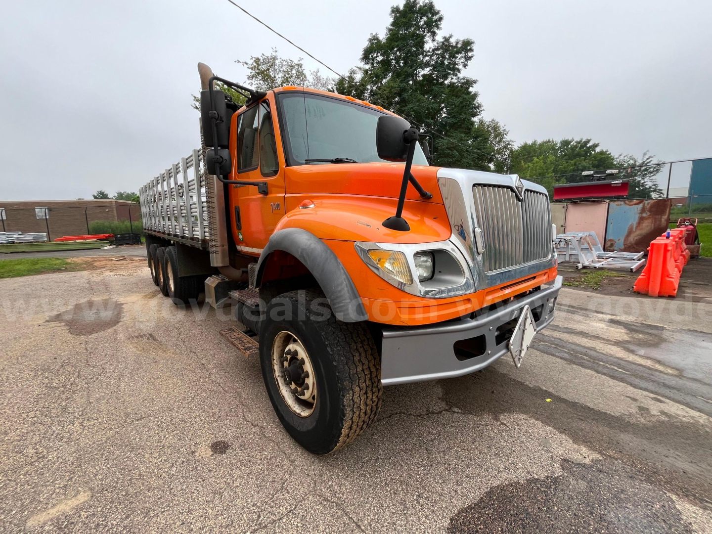 Orange 2004 International 7600 flatbed dump truck, front view with chrome grille and bumper, large tires, white stakes in bed, overcast sky.