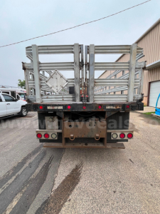 Back of flatbed dump truck with metal gating and dual taillights. Visible text includes a diamond placard holder on the gate and taillight cluster.