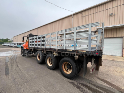 2004 International 7600 flatbed dump truck with long dual rear axles and metal stake sides, orange cab, parked outside a warehouse.