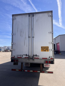 Rear view of a utility trailer with double doors, metal exterior, and caution sticker on the right door. Flatbed framed bumper with red reflectors.