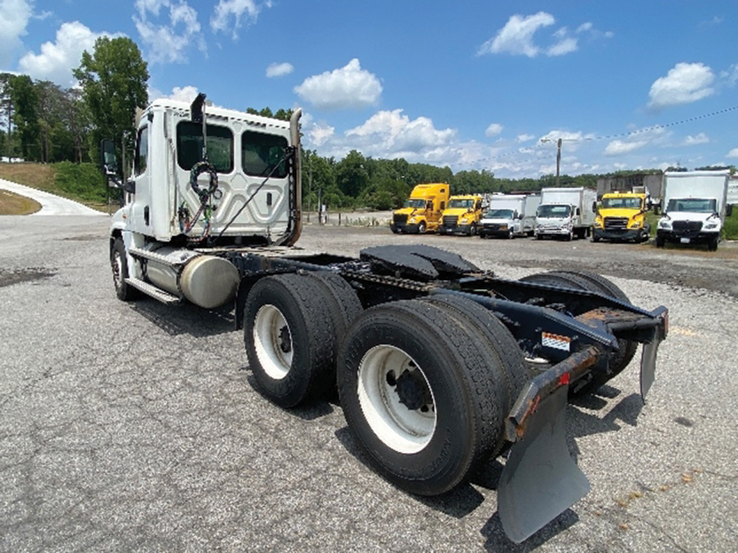 Truck chassis and cab of a 2020 Freightliner Cascadia125 tractor, rear dual axles visible with white rims, fifth-wheel and mud flaps; yard with other trucks in background.