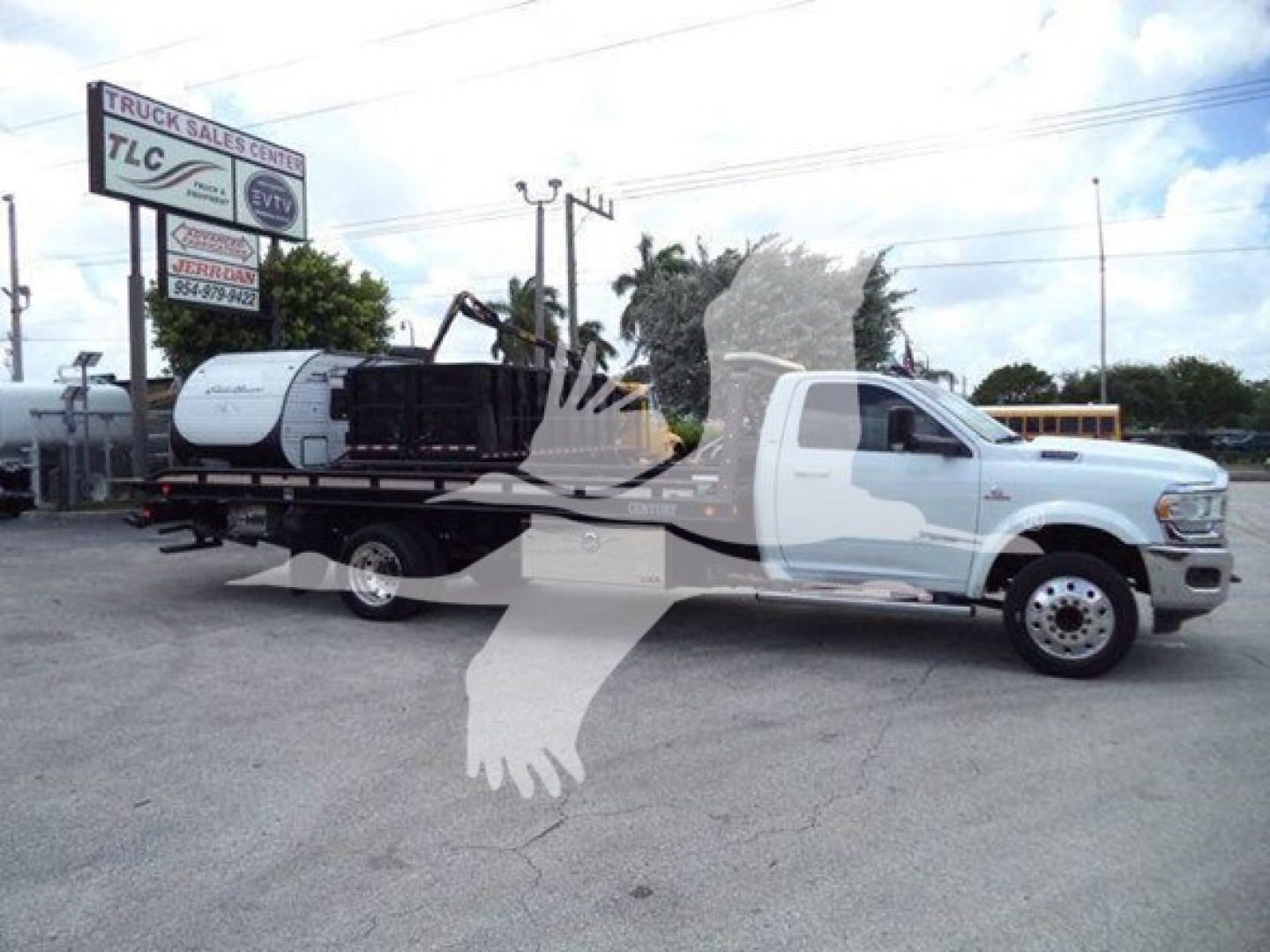 Ram 5500 rollback tow truck with white cab and flatbed carrying a compact excavator; dealership lot in background with TCL sign.
