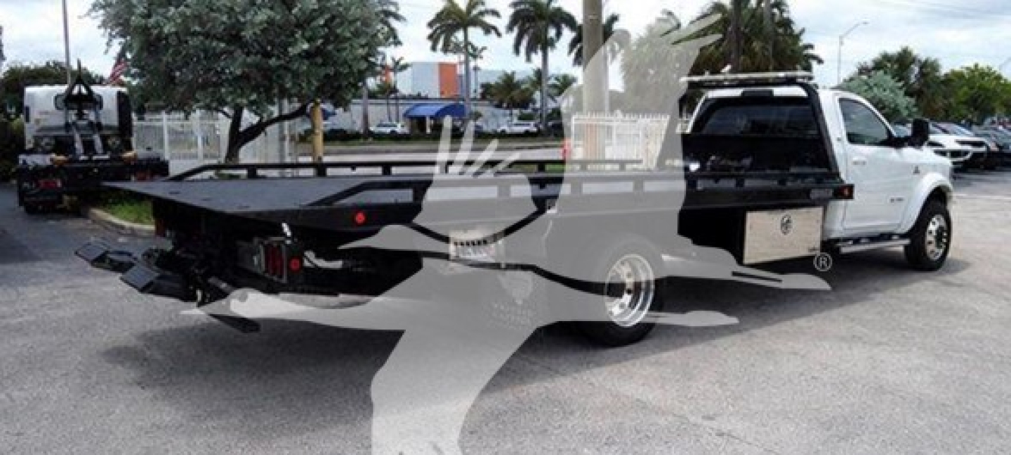 Flatbed rollback tow truck with a white cab and black flatbed, viewing left side and rear, showing truck bed and equipment rails.