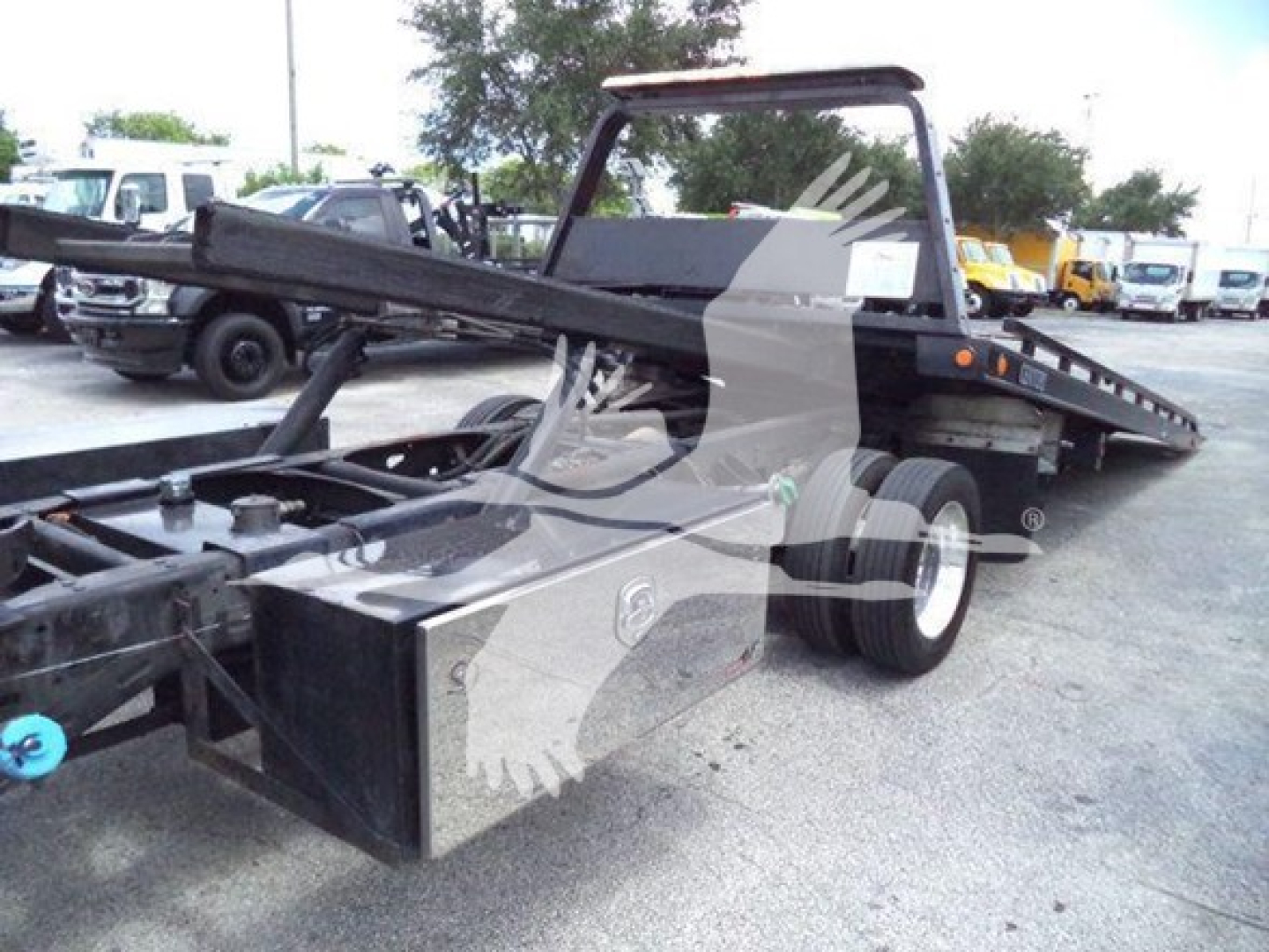 Truck flatbed rollback with hydraulic tow line showing metal frame, dual rear wheels, and a front toolbox. Visible text on components none legible.