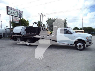 Ram 5500 rollback tow truck with white cab and flatbed carrying a compact excavator; dealership lot in background with TCL sign.