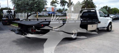 Flatbed rollback tow truck with a white cab and black flatbed, viewing left side and rear, showing truck bed and equipment rails.