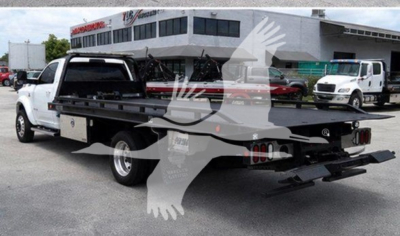 Ram rollback tow truck in yard showing flatbed with wheel lift and ramps; dealership building in background.