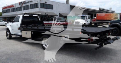 Truck rollback equipment on trailer showing flatbed with branding and background dealership building.