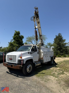 Truck-mounted digger/derrick unit on a white GMC C8500 utility truck, with tall drilling rig and auger visible, front bumper and grille, daytime outdoor lot.