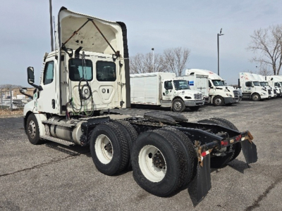 Truck chassis with cab raised showing Freightliner Cascadia driveline and dual rear axles; background includes a row of white delivery trucks.