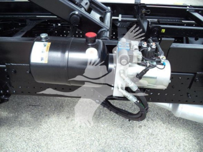 Truck component showing air tank, hydraulic pump, and control assembly mounted to a frame with hoses and wiring visible.