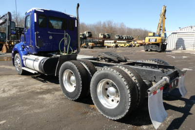 Blue Kenworth truck cab with twin rear axles and mud flaps, parked in a lot with construction equipment in background.
