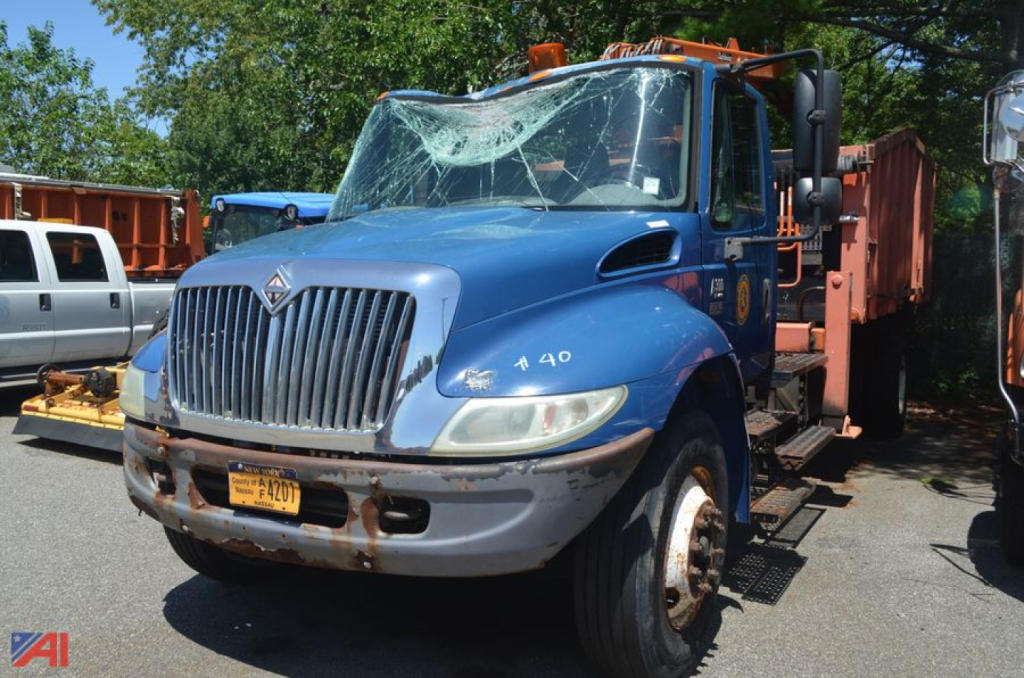 Truck front view of a blue 2005 International DuraStar 4300 loader with broken windshield and orange equipment on flatbed, license plate visible