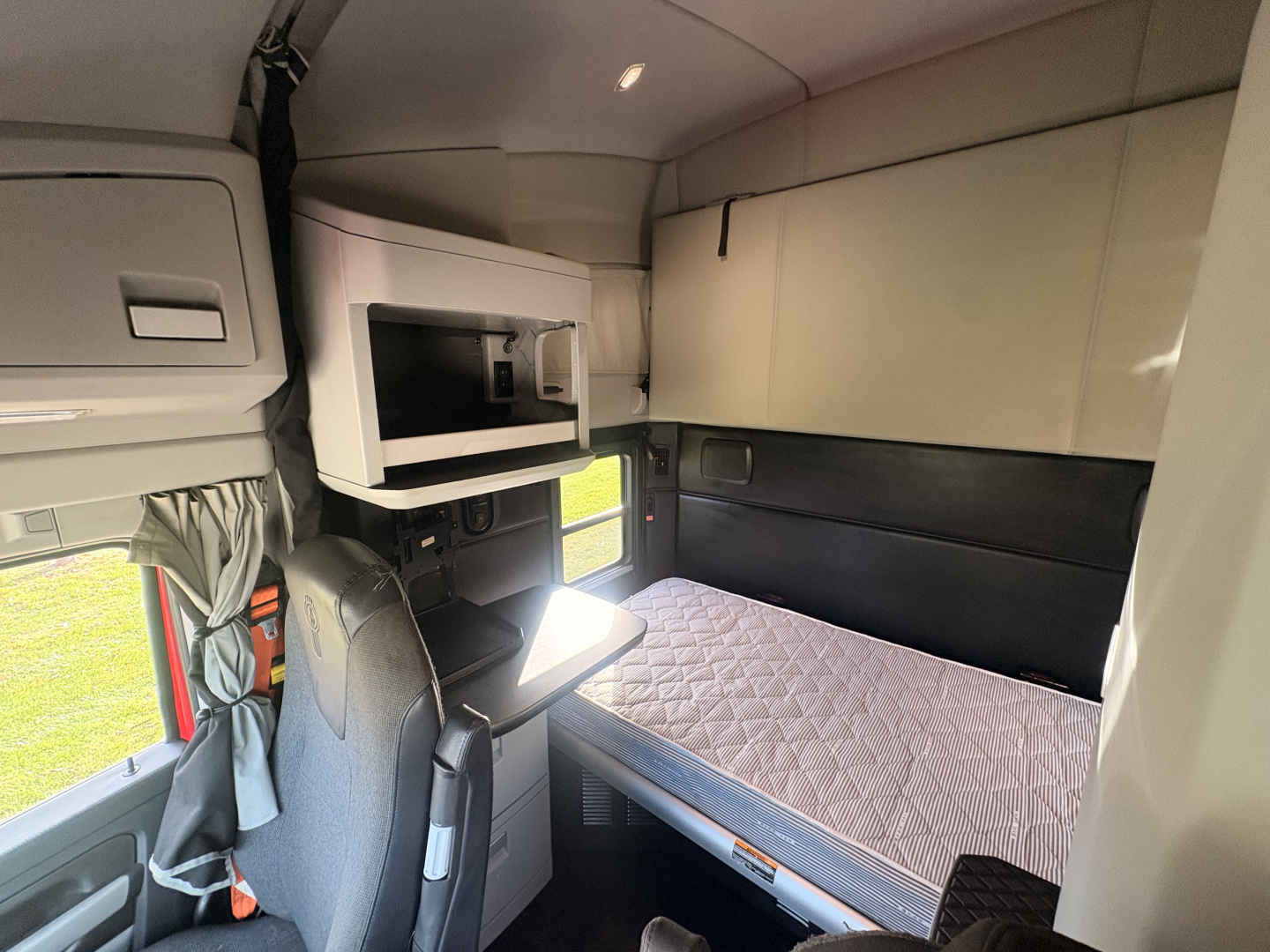 Kenworth T680 cab interior with sleeper bunk, upper storage cabinets, mounted microwave, and window view.