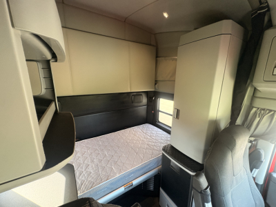 Kenworth T680 cab interior showing sleeper bunk with quilted mattress, overhead storage cabinets, and a refrigerator unit near the window.
