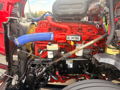 Engine bay of a 2023 Kenworth T680 showing bright red engine with blue hose, intake manifold labeled XDIS, radiator area, and various hoses, wires, and a white reservoir.