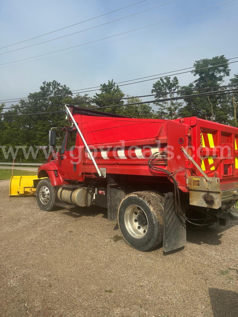 Dump truck with red hopper and plow attachment at front, side view showing dual rear tires and hydraulic hoses.