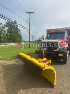 Heavy-duty plow attached to a red International truck in a roadside setting, with a large yellow blade and hydraulic mounting.
