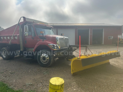Truck with a yellow snow plow attachment mounted to the front, red International 7400 cab, snow plow wide blade extending to the right, in an outdoor yard.